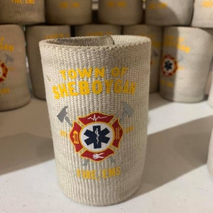 May include: A white fabric can cooler with the text "Town of Sheboygan" in yellow. The cooler has a red and yellow fire department logo with a star of life and the words "Fire/EMS".