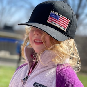 May include: A young girl wearing a black and white baseball cap with a red, white, and blue American flag patch that says "Crafty American". She is also wearing a purple and white fleece jacket with a Columbia logo.