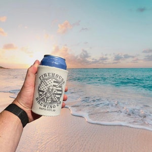 May include: A white can cooler with the text "Firehouse Brewing Co. Rapid City, SD" printed on it. The cooler is holding a blue can of beer. The can cooler is being held by a hand in front of a beach scene with blue water and white sand.