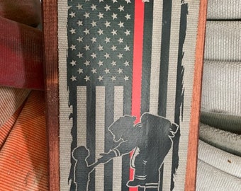 May include: A wooden sign with a red, white, and blue American flag with a thin red line running down the center. The flag is printed on a canvas-like material. A black silhouette of a firefighter holding a child is in front of the flag. The sign has a dark wood frame with brass-colored screws.
