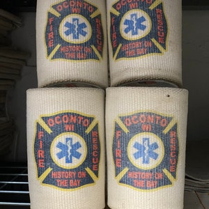 May include: Four white canvas fire hose holders with the Oconto, WI Fire & Rescue logo. The logo features a red cross with a blue star of life in the center. The text "History on the Bay" is printed below the star of life.