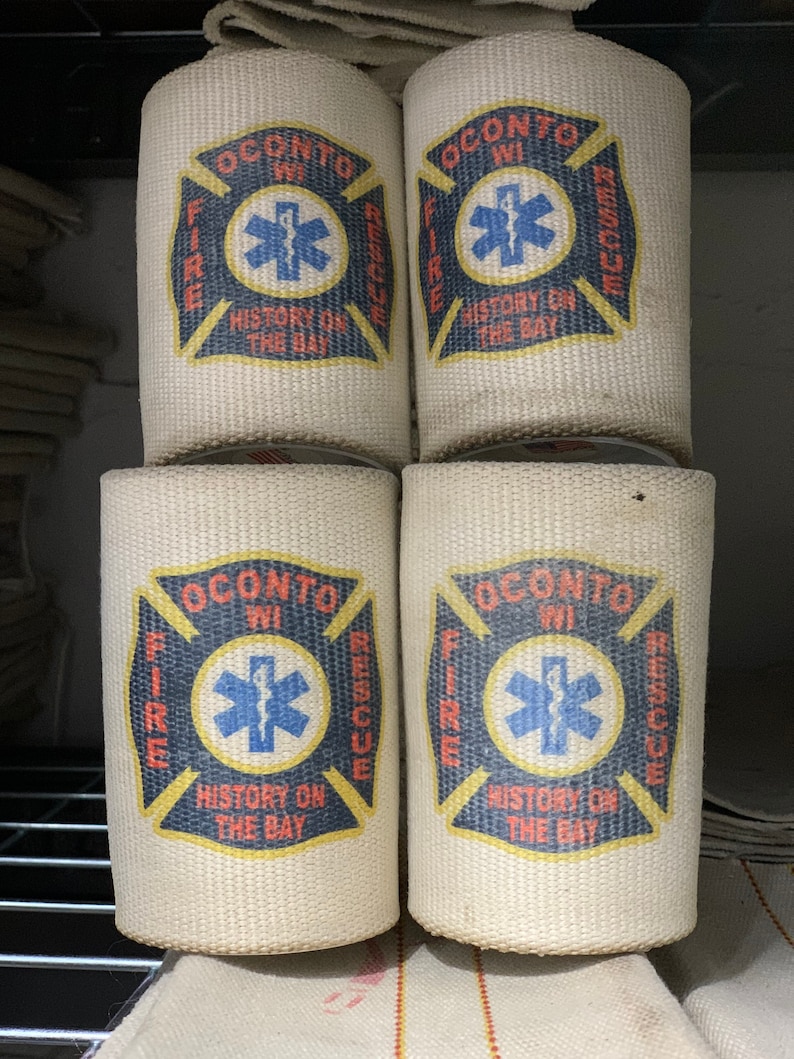 May include: Four white canvas fire hose holders with the Oconto, WI Fire & Rescue logo. The logo features a red cross with a blue star of life in the center. The text "History on the Bay" is printed below the star of life.