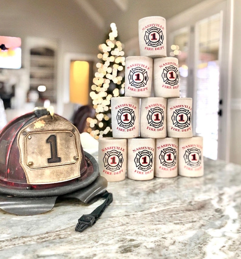 May include: A stack of white can coolers with the words "Nashville Fire Dept" and "Engine 1" printed in red. The can coolers are arranged in a pyramid shape. A red and white fire helmet with a large number "1" on the front is in the foreground.