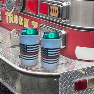 May include: Two blue and white can coolers with a red stripe and an American flag design. The coolers are sitting on the back of a red fire truck.