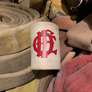 May include: A white canvas fire hose strap with a red embroidered "FC" logo. The strap is surrounded by rolled up fire hoses.