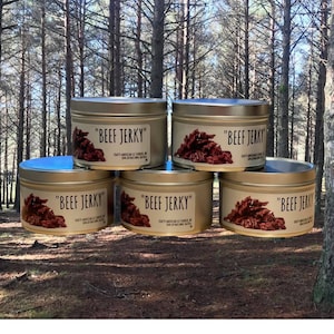 Op de afbeelding: Vijf gouden metalen blikken met deksels, elk gevuld met gedroogd rundvlees. De blikken zijn in een piramidevorm gestapeld en hebben een etiket met de tekst "Beef Jerky".