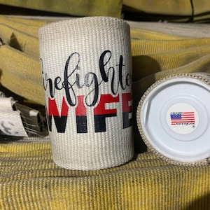 May include: White fire hose fabric can cooler with black and red text that reads "Firefighter Wife". The can cooler has a white lid with a red, white, and blue American flag sticker that reads "Crazy American".