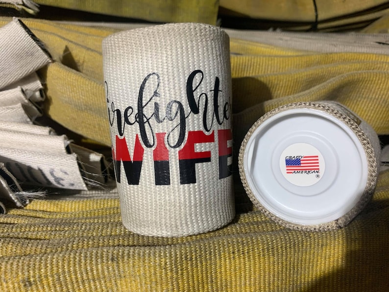 May include: White fire hose fabric can cooler with black and red text that reads "Firefighter Wife". The can cooler has a white lid with a red, white, and blue American flag sticker that reads "Crazy American".