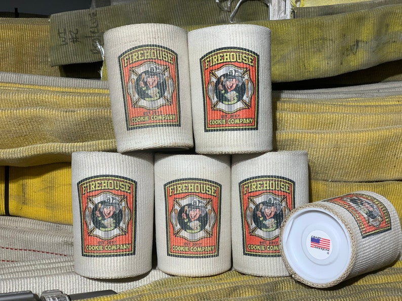 May include: Six white canvas can coolers with a red, yellow, and black logo that says "Firehouse Cookie Company" and a cartoon image of a firefighter. The logo is surrounded by a red border. The can coolers are stacked on top of each other. The background is a yellow fire hose.