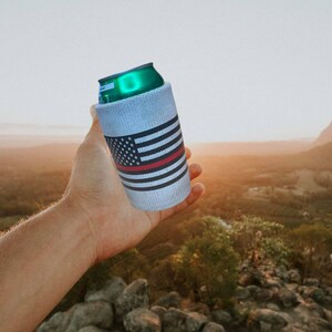 May include: A white can cooler with an American flag design and a red stripe. The cooler is made of a soft, insulated material and is perfect for keeping drinks cold.