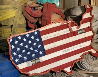 May include: A red, white, and blue American flag made from fire hose, shaped like the United States. The flag is hanging on a wall with fire hoses in the background. The flag has the text "TEST TO 400 PSI PER NFPA 1962" and "PULASKI FIRE DEPT." printed on it.