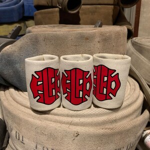 May include: Three white fabric can koozies with a red fire department logo. The koozies are sitting on top of a pile of fire hoses.