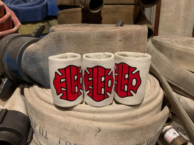 May include: Three white fabric can koozies with a red fire department logo. The koozies are sitting on top of a pile of fire hoses.