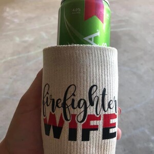 May include: A white fabric can cooler with the text "firefighter wife" in black and red lettering. The can cooler is holding a green and red can of beer.