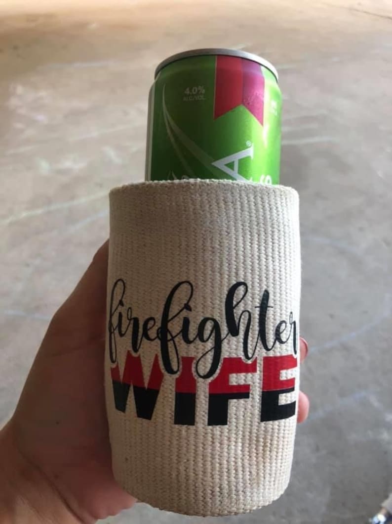 May include: A white fabric can cooler with the text "firefighter wife" in black and red lettering. The can cooler is holding a green and red can of beer.