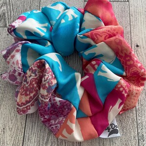 May include: A colorful scrunchie with a mix of patterns and colors, including blue, pink, and white. The scrunchie is made of a silky material and has a tag that says "y De".