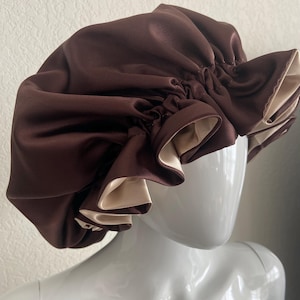 May include: A brown satin bonnet with a contrasting beige interior. The bonnet is gathered at the top and has a ruffled edge. The satin material gives it a smooth, shiny appearance. The bonnet is designed to protect hair.
