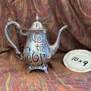 May include: A silver-toned metal teapot featuring an ornate handle, a curved spout, and three decorative feet. The body of the teapot is inscribed with the black text "We the People". Beside it sits a small white plate with "10 x 9" written on it.