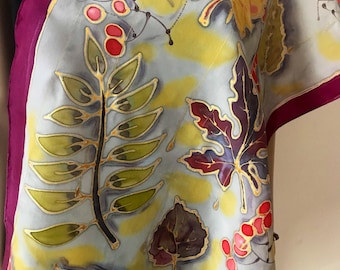 May include: A silk scarf with a botanical design. The scarf features a light blue background with yellow and gold accents. The design includes green, purple, and yellow leaves, along with red berries. The scarf is trimmed with a deep purple border.