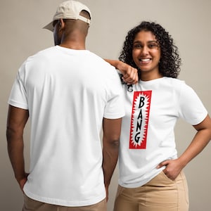 Könnte beinhalten: Zwei Personen tragen weiße T-Shirts. Auf einem Shirt steht das Wort "BANG" in schwarzen Buchstaben in einem roten Rechteck mit weißem Zackenrand. Die andere Person trägt ein schlichtes weißes T-Shirt und eine beige Baseballkappe.
