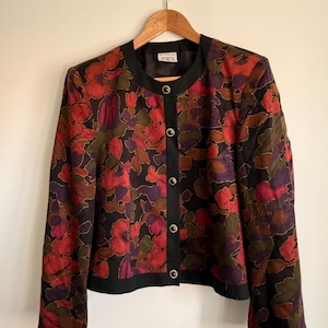 May include: A long-sleeved jacket featuring a black base with a vibrant floral pattern in shades of red, purple, and brown. The jacket has a round neckline, a solid black front placket with five gold-rimmed buttons, and black trim at the cuffs and hem.