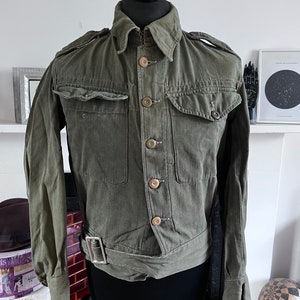 以下が含まれることがあります： ボタン留めのフロント、2つの胸ポケット、ウエストベルトが付いた、オリーブグリーンのミリタリースタイルのジャケット。 ジャケットは襟付きで、肩にエポレットが付いています。 袖口はボタン留めです。 ジャケットは黒いマネキンに展示されています。