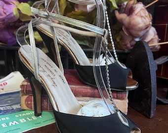 May include: A pair of black satin peep-toe heels with gold trim and a delicate silver ankle strap. The heels have a slender stiletto heel and a decorative chain detail. The shoes are labeled "Jerry de Fourte, London, Made in England."