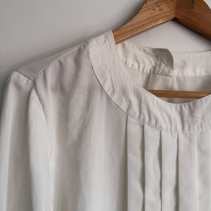 May include: A solid white long-sleeved blouse featuring a round neckline with vertical pleats extending from the collar. The garment is constructed from a smooth, lightweight fabric and is displayed on a wooden hanger against a plain wall.