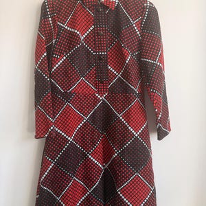 Puede incluir: Un vestido de manga larga con un llamativo patrón geométrico de cuadrados rojos y negros separados por líneas de puntos blancas. El vestido tiene un cuello puntiagudo, una tapeta frontal con botones oscuros y un corte estructurado.