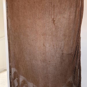 May include: A brown crochet shawl featuring an open-knit mesh pattern throughout the body. The lower section displays a decorative border with repeating oval-shaped motifs, finished with long, thin fringe tassels along the bottom edge.