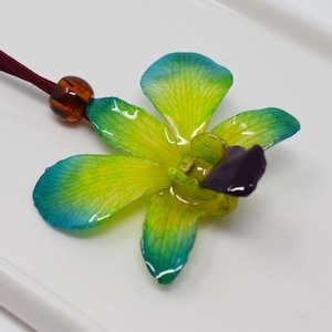 Puede incluir: Una flor de orquídea amarilla con detalles verdes y azules, hecha de resina, cuelga de un cordón marrón con una cuenta marrón. La flor es un collar con colgante.