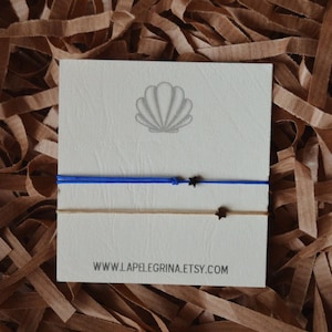 May include: Two friendship bracelets with black star charms. The top bracelet is blue and the bottom bracelet is beige. The bracelets are displayed on a white card with a seashell graphic and the text "www.lapelegrina.etsy.com".