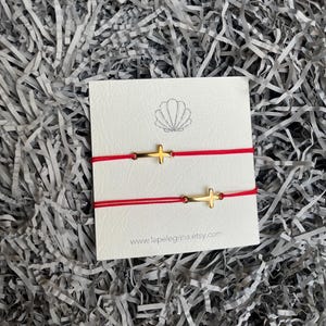 May include: Two red string bracelets with gold cross charms. The bracelets are on a white card with a seashell logo and the text "www.lapelegrina.etsy.com".