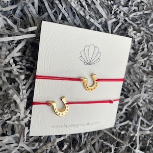 May include: Two red string bracelets with gold horseshoe charms. The bracelets are on a white card with the text "www.lapelegrina.etsy.com".