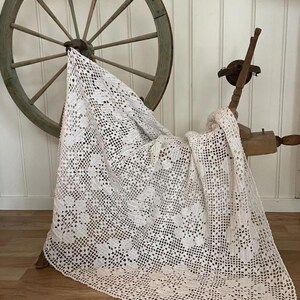 May include: A white, intricately crocheted blanket draped over a wooden spinning wheel and a wooden chair. The blanket features a floral pattern and a delicate, open weave. The background includes a wooden wheel and a white paneled wall.
