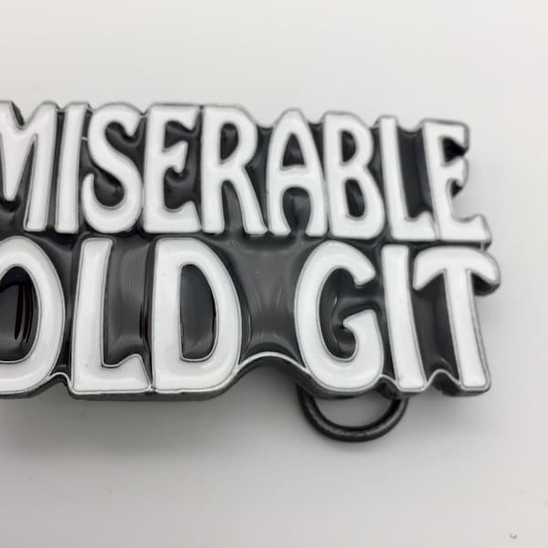 Könnte beinhalten: Rechteckige Gürtelschnalle mit den Worten "MISERABLE OLD GIT" in weißen, erhabenen Buchstaben auf schwarzem Hintergrund. Die Schnalle hat eine silberfarbene Schlaufe zum Befestigen am Gürtel. Ein auffälliges Accessoire.