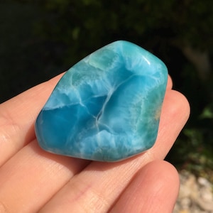 May include: A polished, rounded gemstone with a vibrant blue and turquoise color palette. The stone has a smooth, glossy surface with intricate patterns and veining, resembling ocean waves. The gemstone is held in a hand, showcasing its natural beauty.