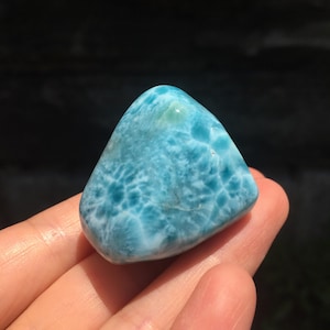 Puede incluir: Una gema pulida de forma triangular, de color azul claro, con intrincadas vetas blancas y azul oscuro. La piedra se sostiene en una mano, mostrando sus bordes redondeados y sus patrones naturales.