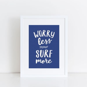 Op de afbeelding: Witte ingelijste print met een blauwe achtergrond en witte tekst die "Worry less Surf more" luidt.