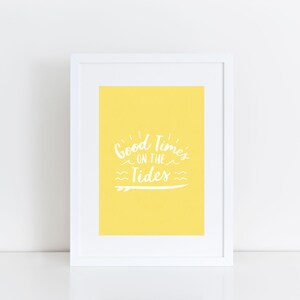 Op de afbeelding: Een wit ingelijst print met een gele achtergrond en de tekst "Good Times on the Tides" in witte letters. De tekst is gestileerd met een surfplank-afbeelding.