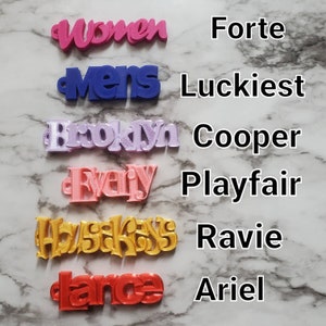 Op de afbeelding: Een verzameling kleurrijke 3D-geprinte woordbedels. De woorden zijn "Women" in roze, "Mens" in blauw, "Brooklyn" in lavendel, "Evany" in perzik, "AbyssKiss" in goud en "dance" in rood. Elke bedel heeft een kleine lus.