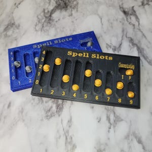 Peut inclure: Deux compteurs de emplacements de sorts en plastique noir et bleu avec des marqueurs dorés. Les compteurs ont les numéros 0-4 et 1-9 imprimés dessus. Le texte "Spell Slots" et "Concentration" est imprimé sur les compteurs.