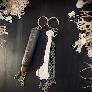 May include: Two unique earrings. One is a dark gray cylinder with gold markings and chain fringe. The other is a white bone with chain fringe, both suspended from gold-colored hooks. The earrings are displayed on a dark surface.
