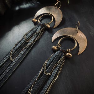 May include: A pair of handcrafted earrings featuring crescent moon-shaped pendants in a bronze finish. Each earring has a circular ring with small bells and black leather fringe accented with delicate chains. The earrings are designed for pierced ears.