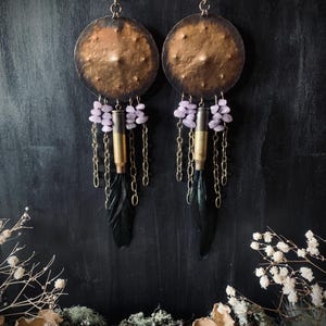 May include: A pair of handcrafted earrings with a bohemian aesthetic. Each earring features a hammered copper disc, small purple beads, brass bullet casings, black feathers, and delicate chain accents. The earrings hang from a simple hook.
