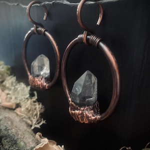 May include: A pair of hoop earrings crafted from dark copper wire, featuring a textured, wrapped base that secures a clear, pointed quartz crystal within the center of each circular frame. The earrings are suspended from a thin, dark wire.