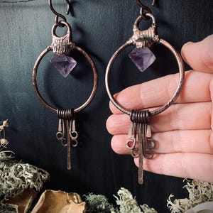 May include: A pair of handcrafted copper earrings. Each earring features a large copper hoop with a faceted purple gemstone and decorative copper wire accents. The earrings have a rustic, bohemian aesthetic.