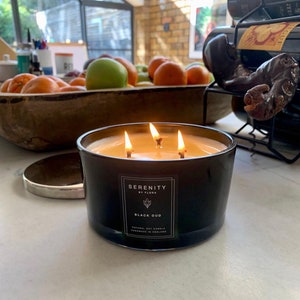 May include: A black glass candle with three wicks burning. The candle is labelled "Serenity by Flora" and "Black Oud". It is a natural soy candle handmade in England.