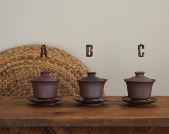 May include: Three brown ceramic teacups with lids and matching saucers. The teacups are arranged in a row on a wooden table. The teacups are labeled A, B, and C.
