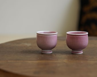 May include: Two small, matching, mauve-colored ceramic cups with a white rim and base sit on a wooden table. The cups have a rounded shape and a footed base. The background is blurred, with a neutral wall and a patterned pillow.